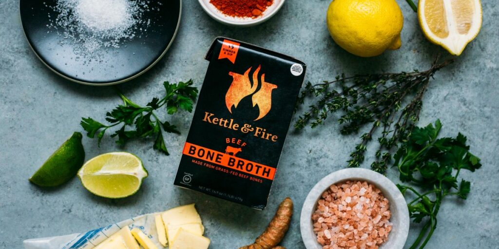 A carton of kettle and fire beef bone broth surrounded by fresh ingredients.