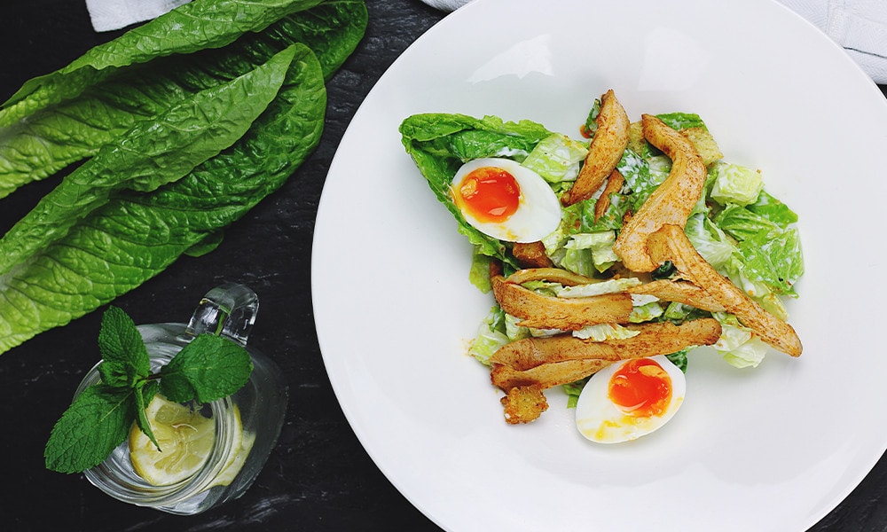 A plate of vegetable salad with an egg that is cut open next to a mason jar with water, a slice of lemon and some mint leaves.