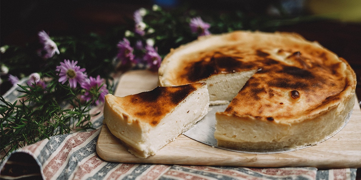 A whole cheesecake with a part that is sliced in triangular shape. Purple flowers at the background.