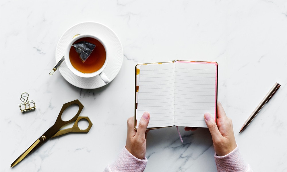 A person holding a blank notebook with pens, pair of scissors, paper clip and a cup of tea on the table.