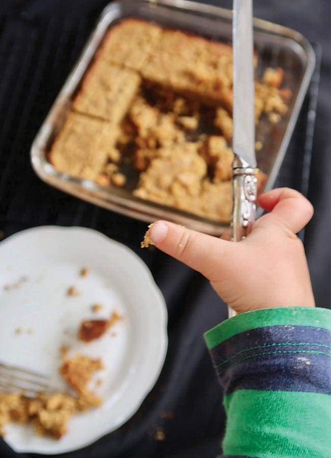 A hand holding a small knife about to take a slice of the cinnamon coffee cake in a baking dish.