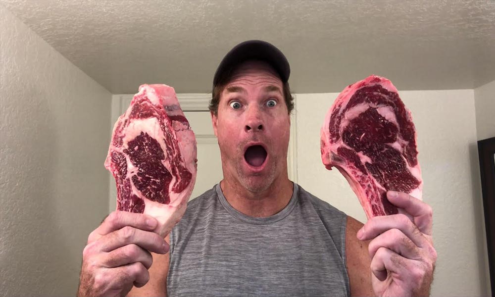 A man is holding up two steaks in front of a mirror.