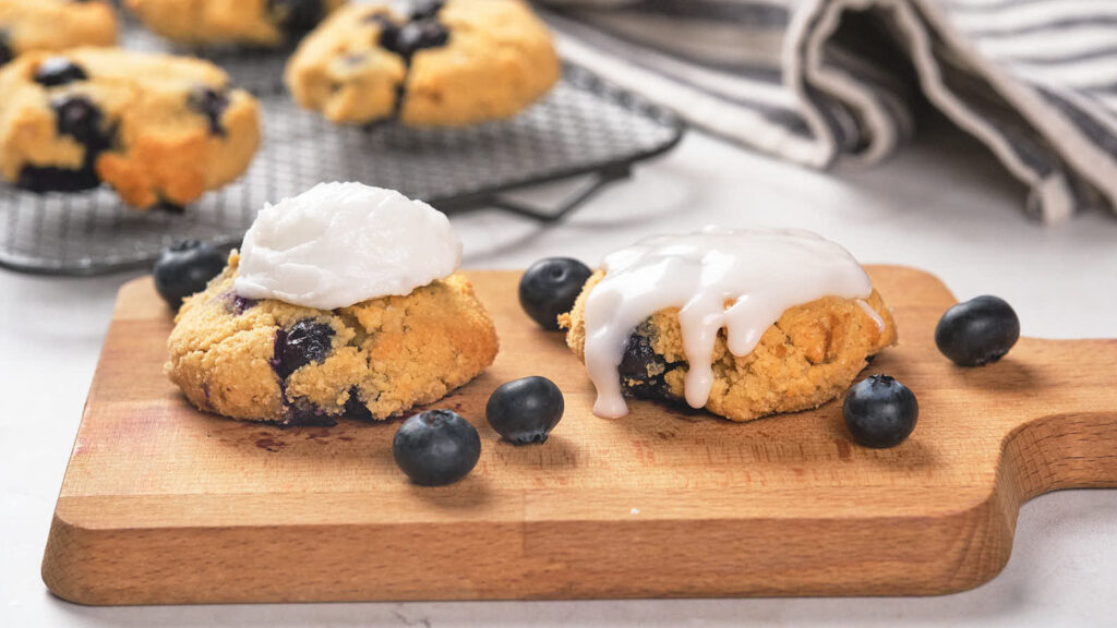 Two blueberry scones on a wooden board, one topped with a dollop of cream and the other with dairy-free keto icing. Fresh blueberries are scattered around them. A cooling rack with more scones is in the background.