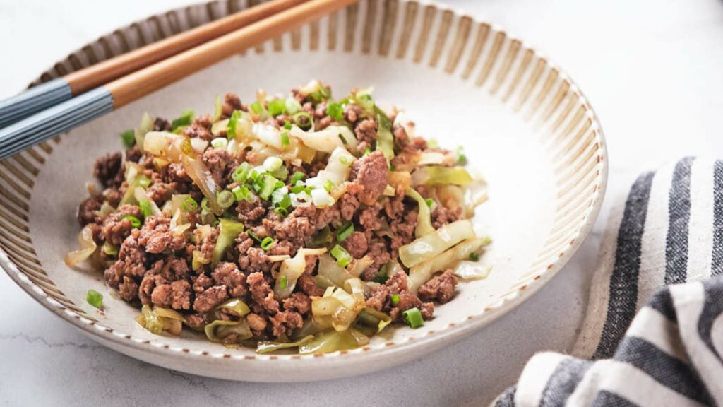 A plate of stir-fried minced meat with chopped green onions and cabbage, served alongside chopsticks on a gray textured surface.