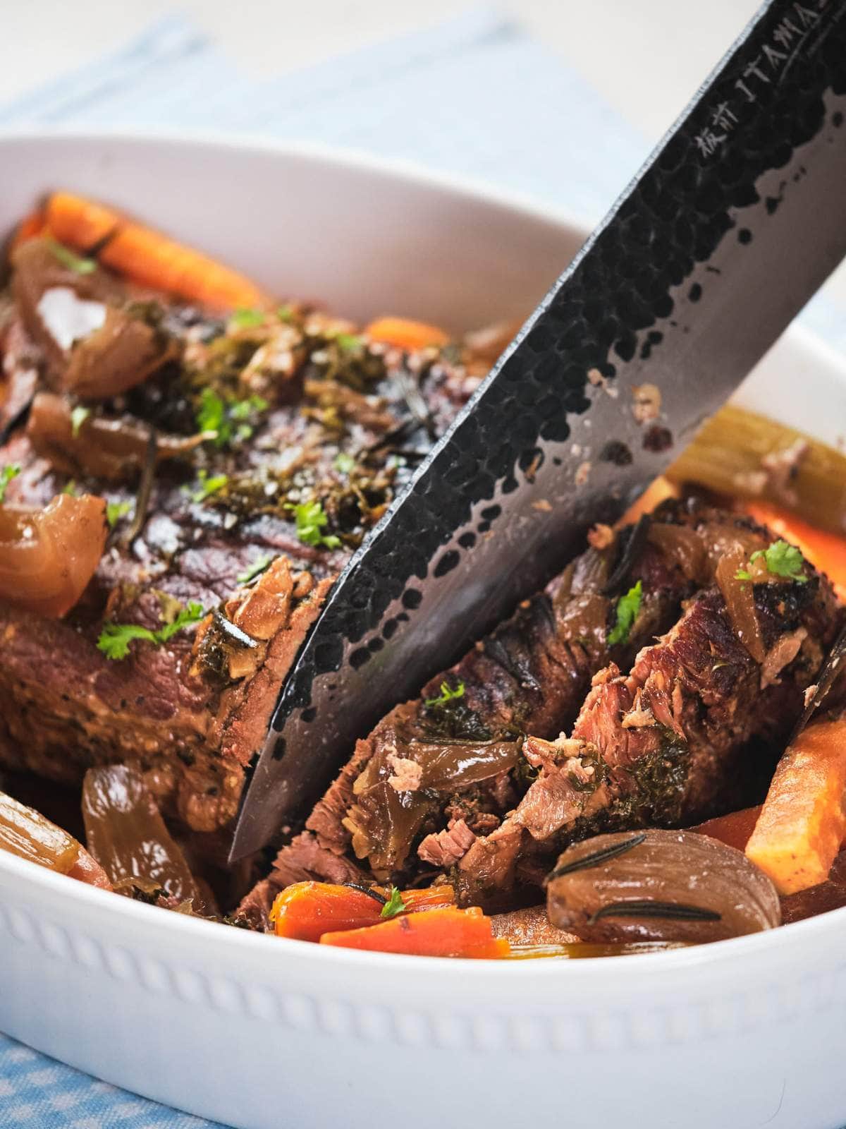 A close-up of keto pot roast with carrots and onions in a white serving dish, garnished with herbs, with a knife resting on top.
