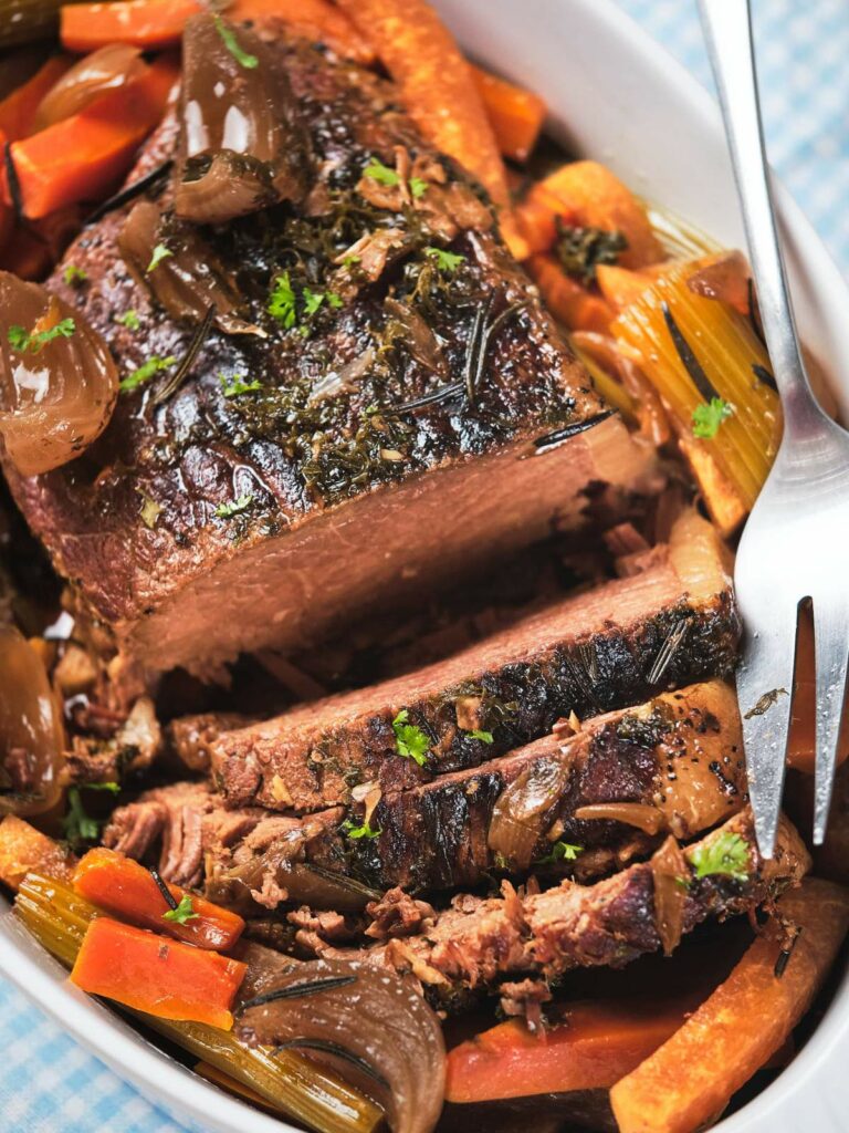 A close-up of a keto pot roast sliced on a plate, garnished with carrots, onions, and herbs.