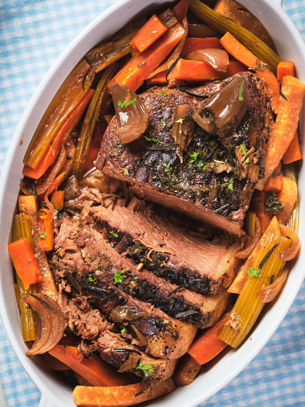 Slow cooker pot roast sliced and served with carrots and onions in an oval dish on a blue checkered tablecloth as a keto pot roast.