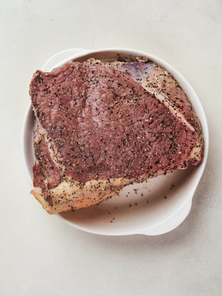 A raw chuck steak seasoned with black pepper and salt on a white plate.