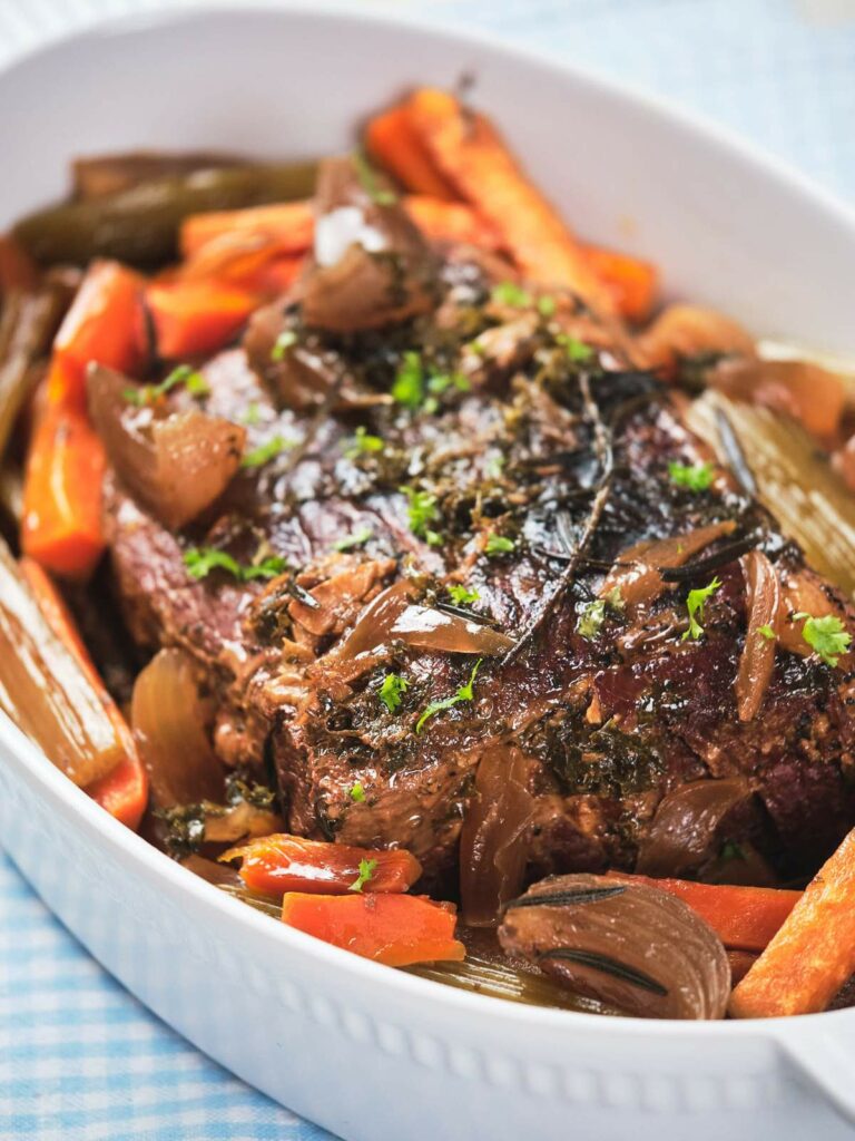 Keto pot roast with carrots, onions, and herbs in a white ceramic dish.