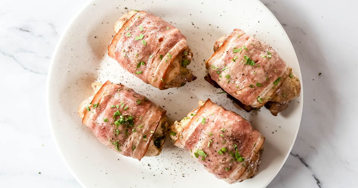 Four bacon-wrapped chicken thighs on a plate with parsley and black pepper garnish.