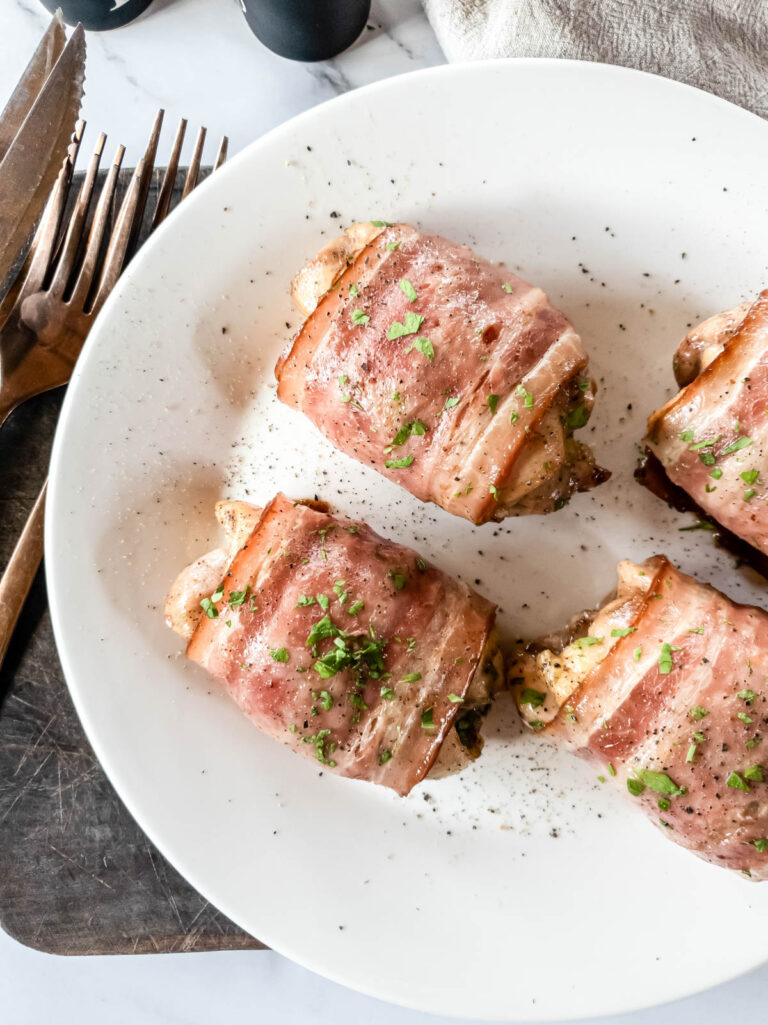 Four chicken thighs wrapped in bacon on plate on cutting board.