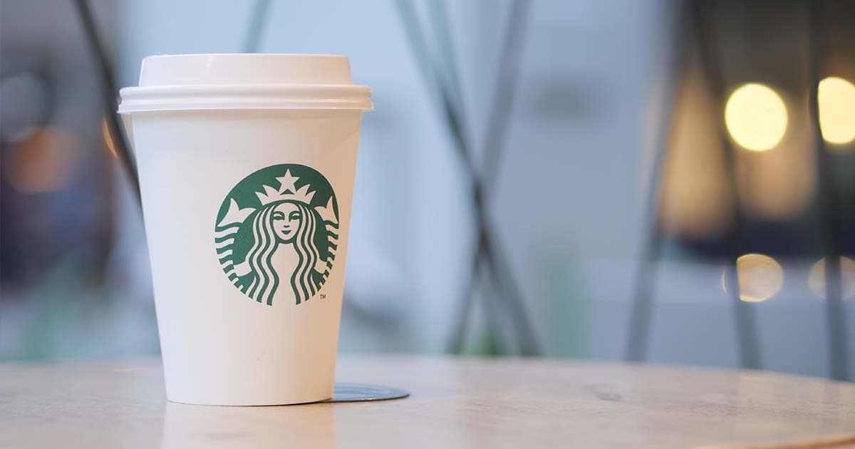 A sugar-free Starbucks coffee cup sits on a table, lights blurred in background.