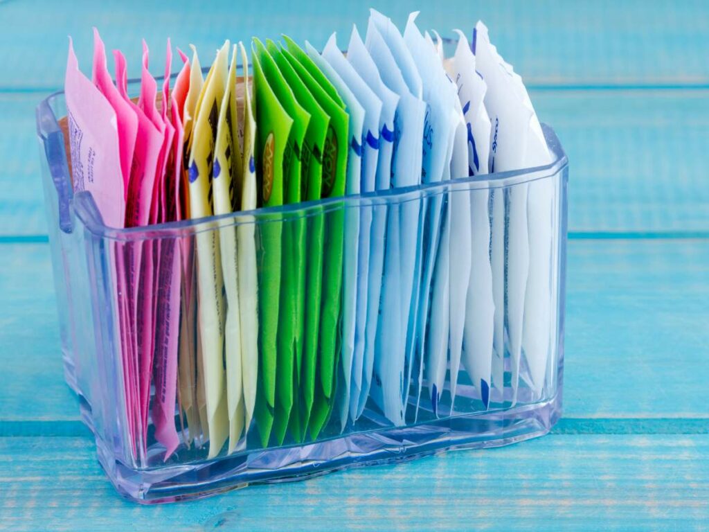 A clear plastic holder containing various colored sugar packets on a blue wooden surface.