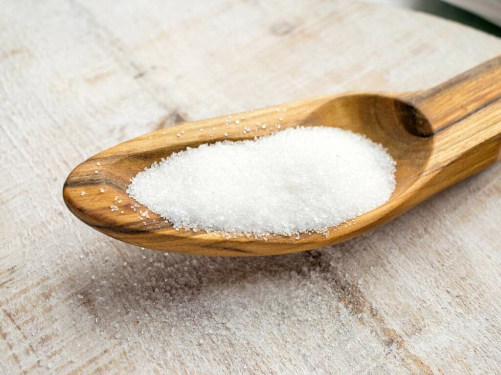 A wooden spoon filled with white granulated sugar on a surface with sugar scattered around.