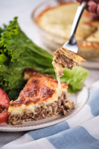 A fork lifting a cheesy slice of meat pie from a plate.