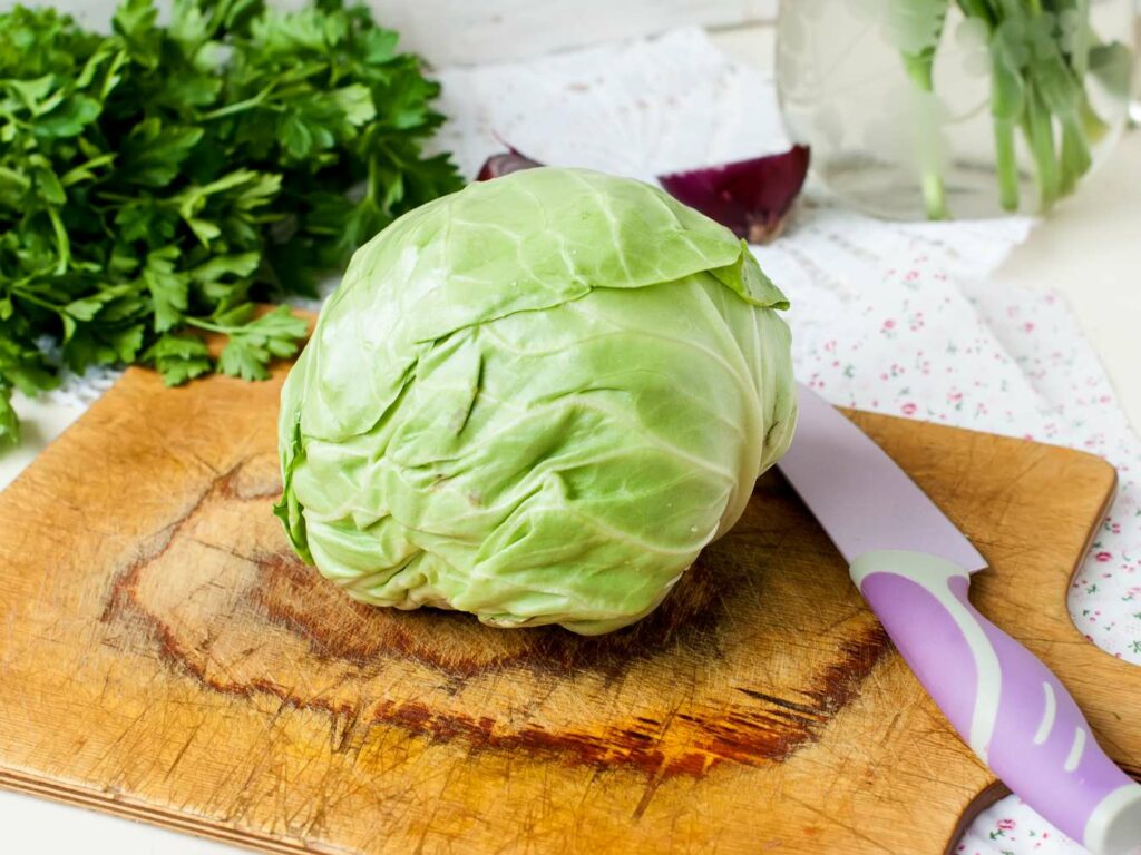 A fresh green cabbage on a wooden cutting board with a purple knife beside it, set against a backdrop of parsley and onions.