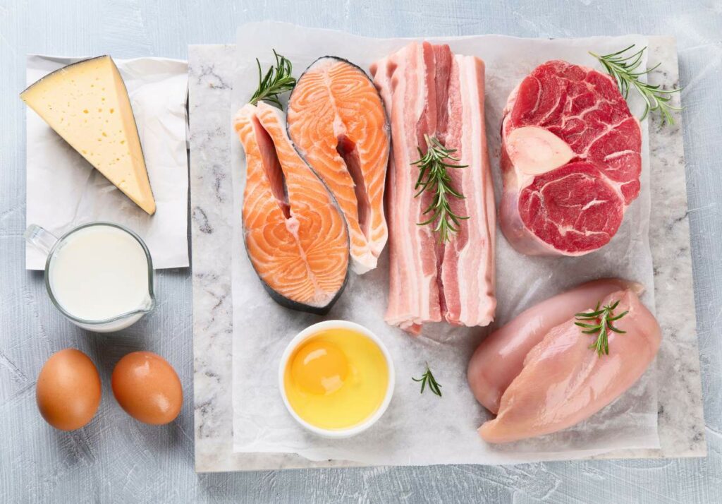 A variety of high-protein foods including cheese, milk, eggs, salmon fillets, bacon strips, a cut of beef, and a chicken breast on a marble surface.