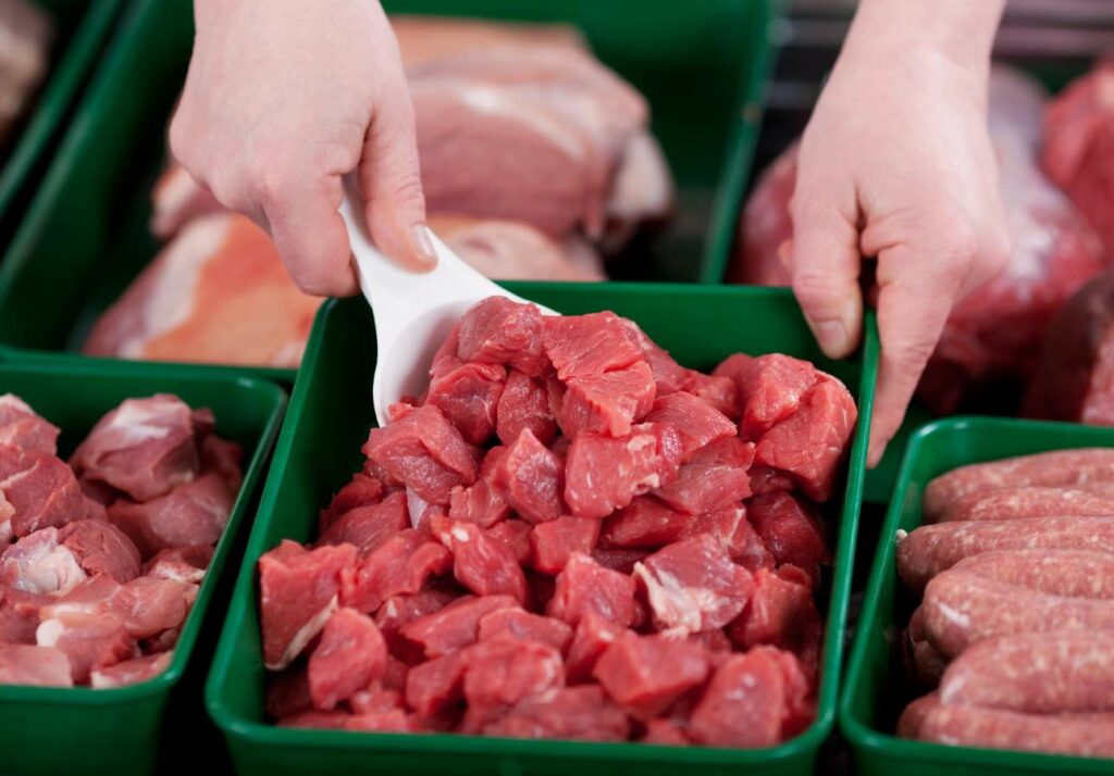 Close-up of hands using a white scoop to pick up pieces of raw red meat from a green container, with other containers of various cuts of meat visible in the background.