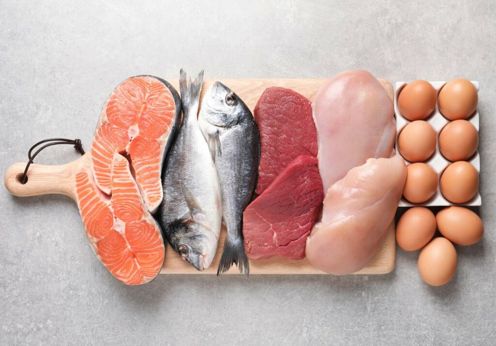 A wooden board with various protein sources: salmon fillets, whole fish, raw steak, chicken breast, and a carton of six eggs, all placed on a light gray surface.