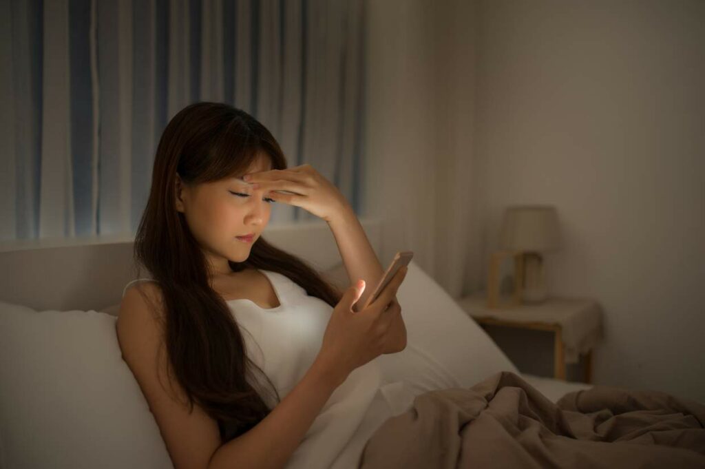 A woman sitting on a bed at night, looking concerned while reading something on her smartphone.