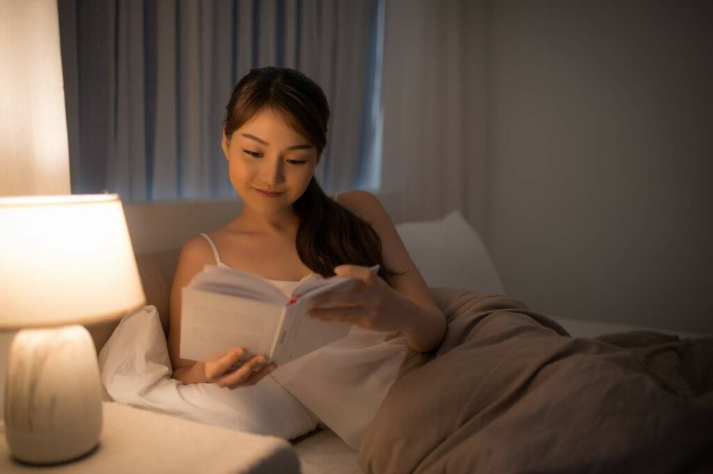 A woman reading a book in bed beside a table lamp at night, with warm lighting in the room.