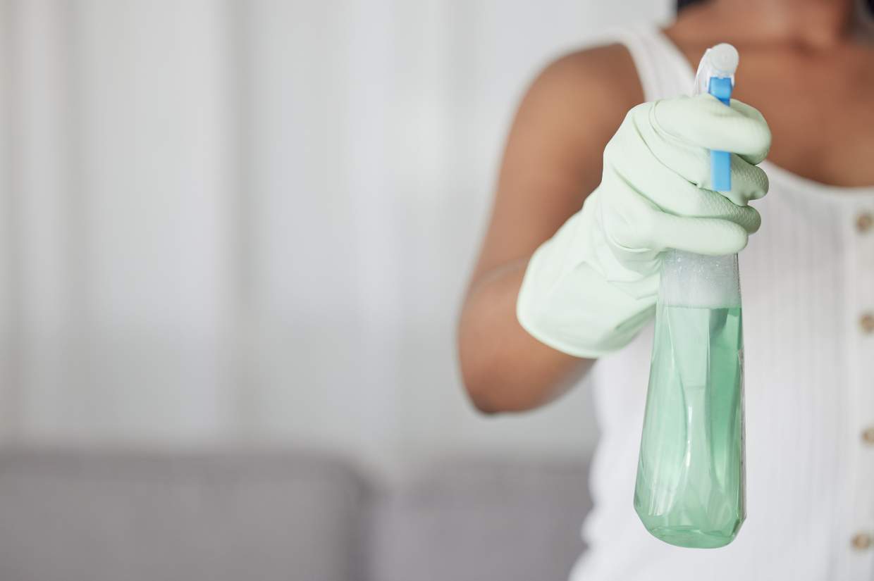 Person wearing a white sleeveless top and green gloves holding a spray bottle with green liquid.