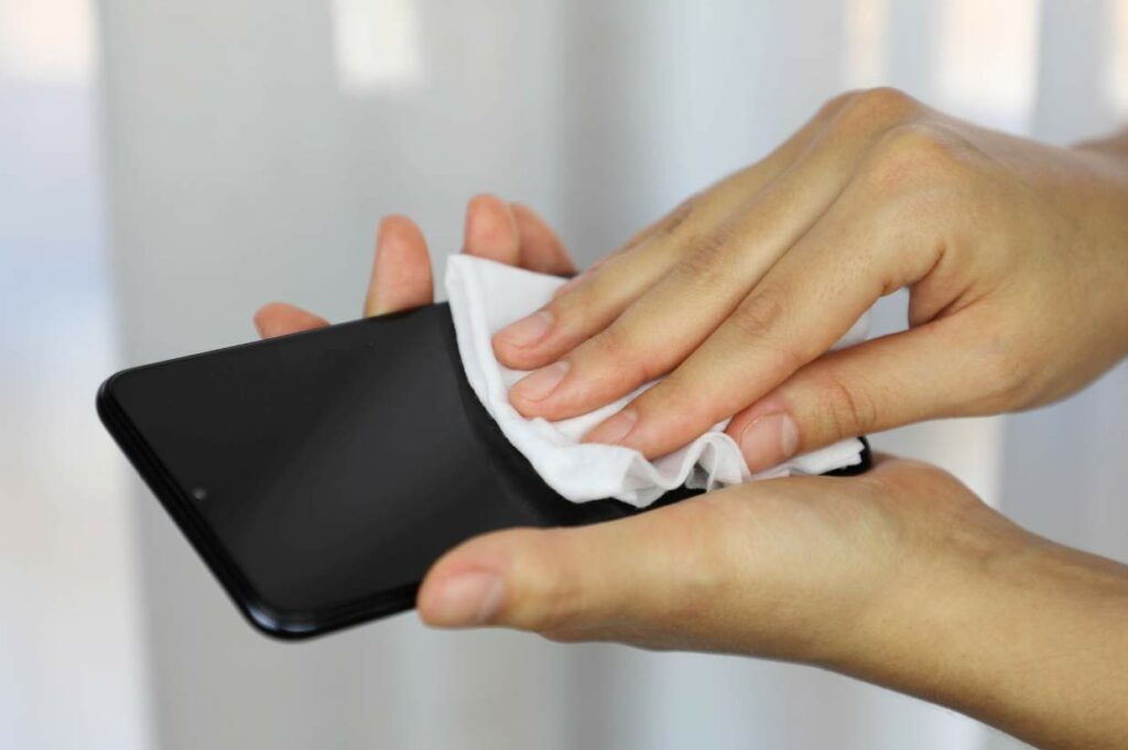 A person gently cleans a smartphone screen with a white cloth against a background of blurred light-colored curtains.