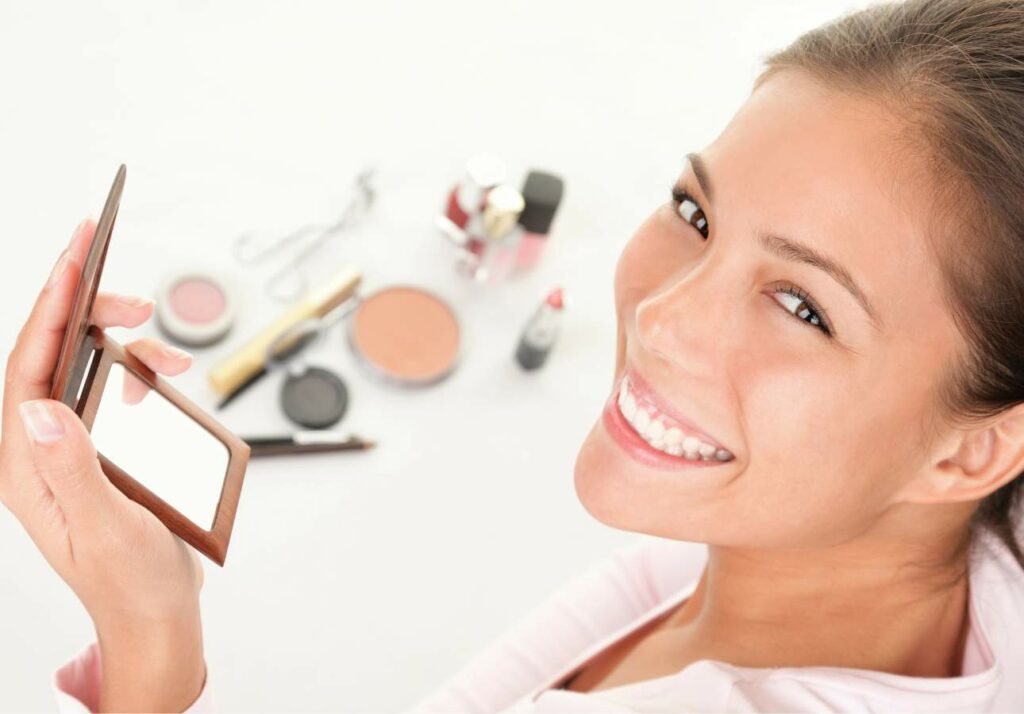 Woman smiling while holding a makeup compact, with various makeup products visible in the background.