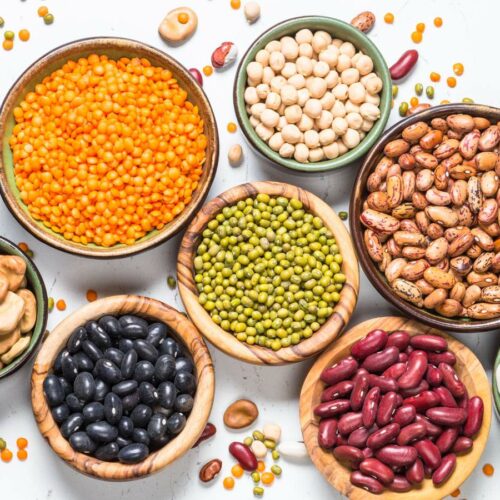 Assorted legumes including lentils, chickpeas, and beans in bowls, spread out on a white background.