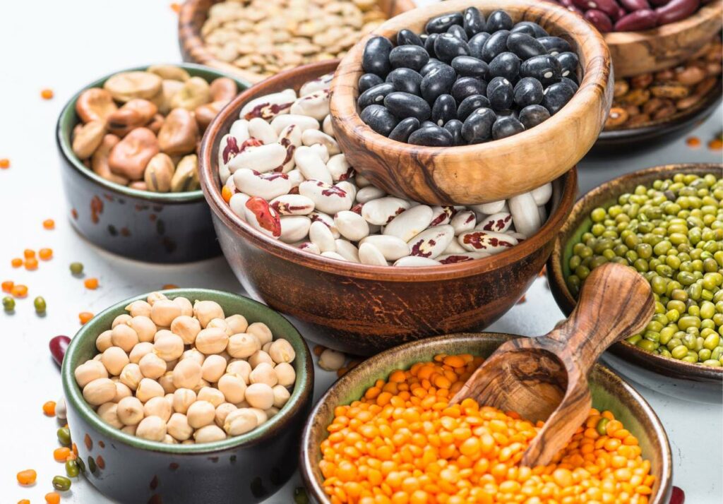 Assorted legumes including black beans, red kidney beans, chickpeas, lentils, and mung beans in wooden and ceramic bowls with a scoop on a white surface.