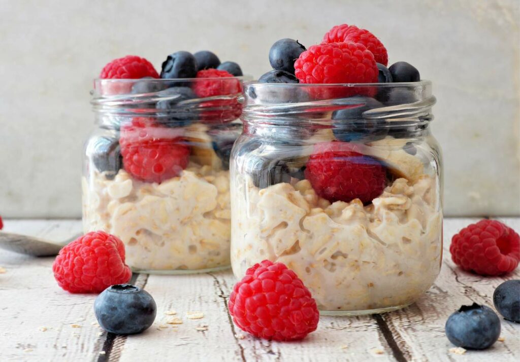 Two glass jars filled with oatmeal and topped with fresh blueberries and raspberries, set on a light wooden surface.