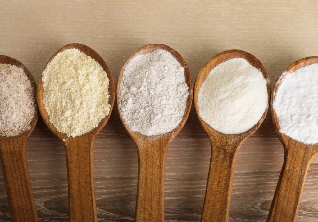 Wooden spoons with different types of flour (corn, wheat, rye, all-purpose) arranged in a row on a wooden surface.