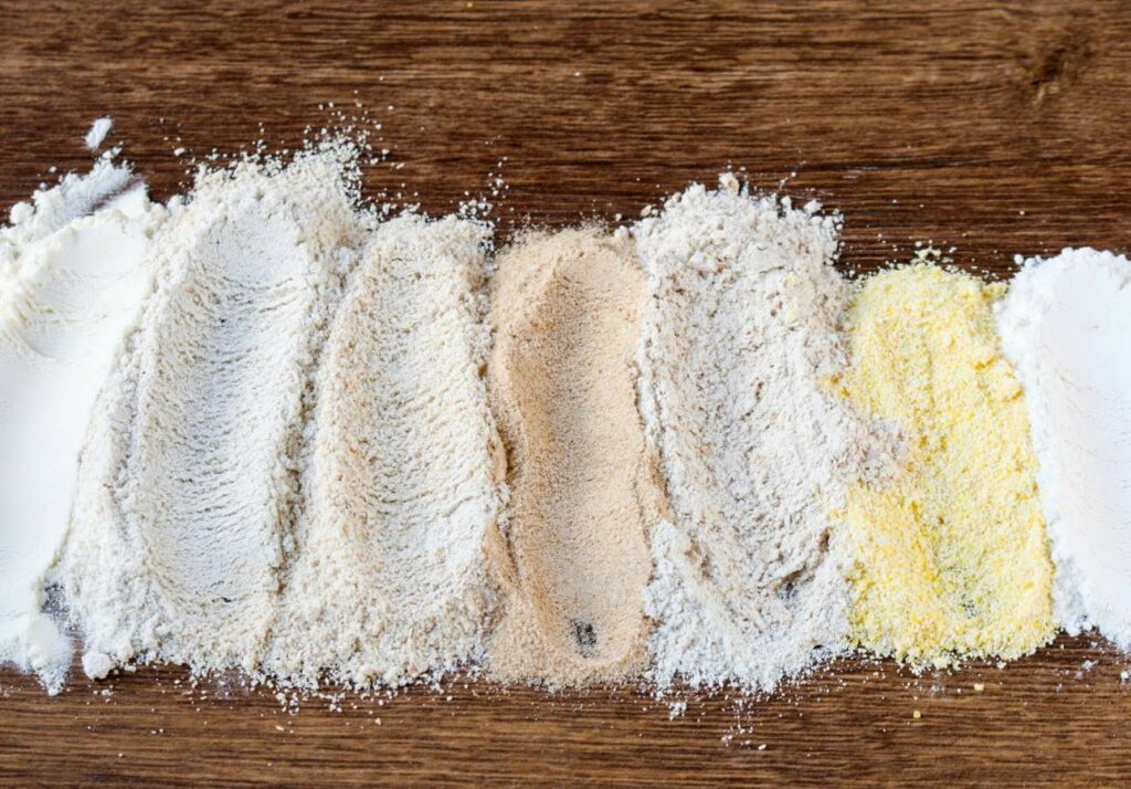 Different types of flour arranged in lines on a wooden surface, showcasing varying textures and colors.