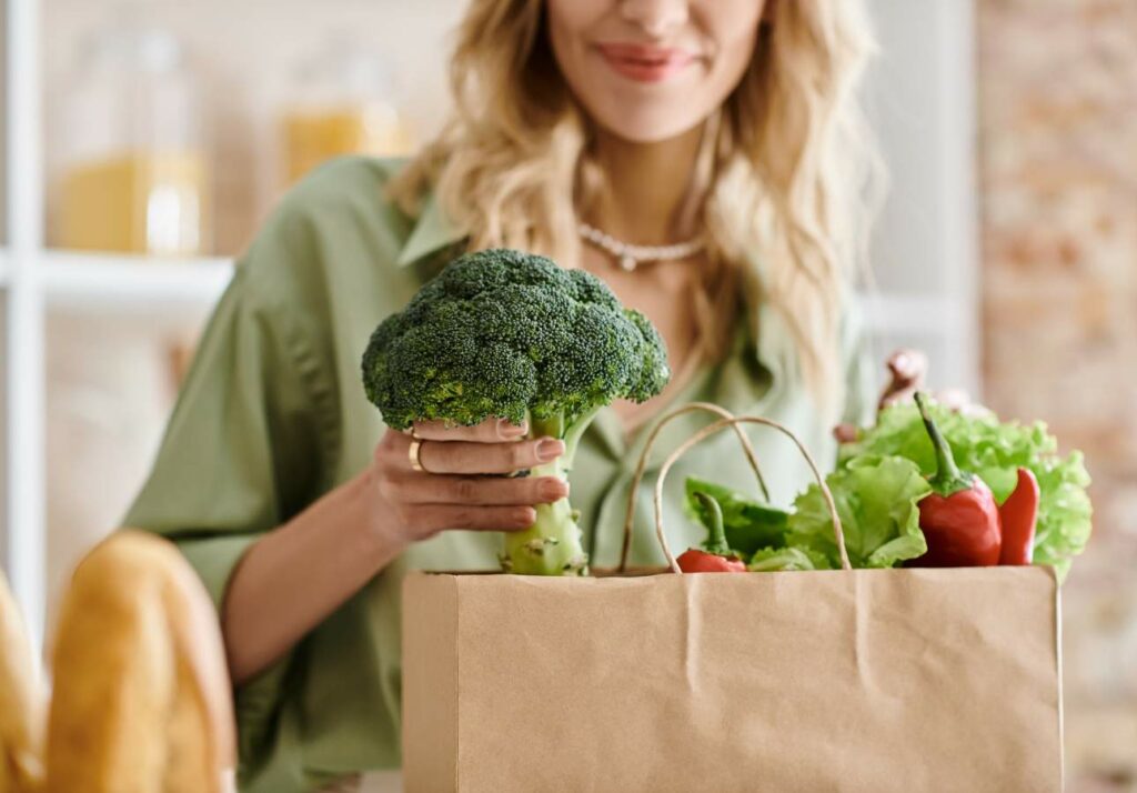 A woman holding a broccoli and unpacking groceries from a brown paper bag filled with fresh vegetables like lettuce and bell peppers in a kitchen.