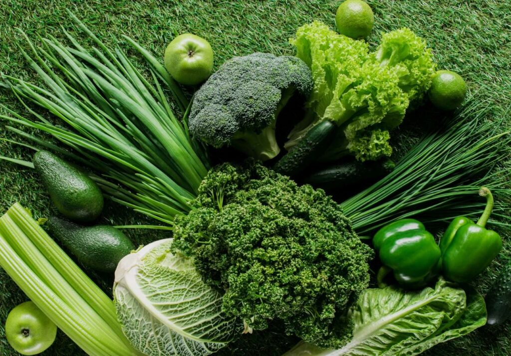 A variety of green vegetables and fruits including broccoli, lettuce, apples, avocados, celery, bell peppers, and leafy greens are arranged on a grassy surface.
