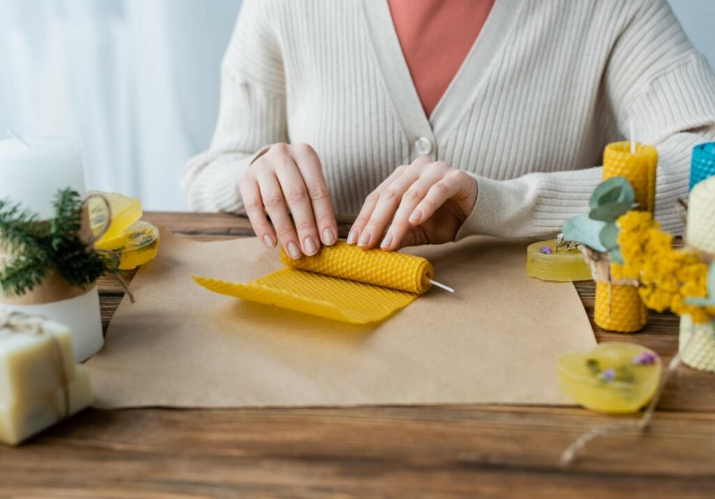 A person is rolling a yellow beeswax sheet into a non-toxic candle at a wooden table surrounded by other crafting materials and finished candles.
