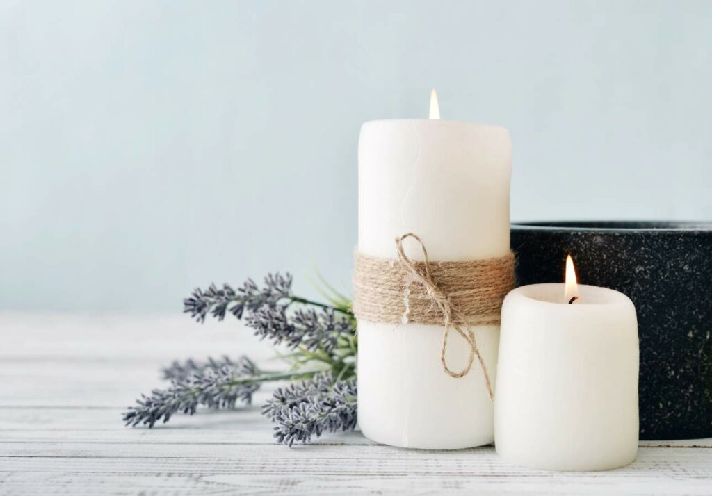 Two white candles, one wrapped with twine, burn on a white wooden surface with lavender sprigs and a black container in the background.
