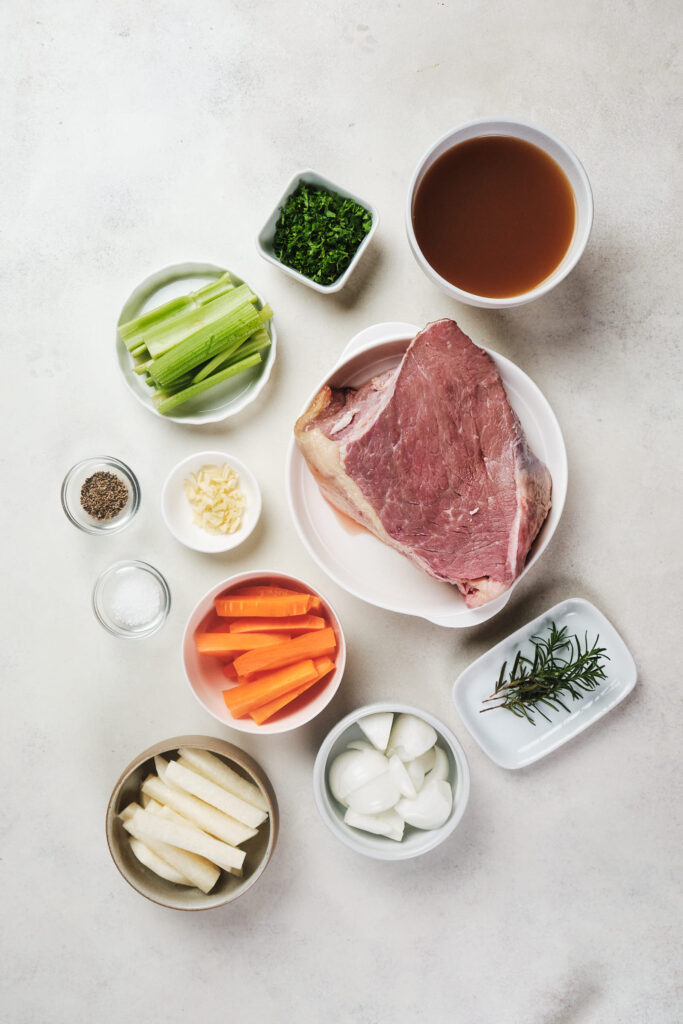 Various ingredients arranged on a table, including a large piece of raw meat, bowls of vegetables like carrots and celery, and seasonings.