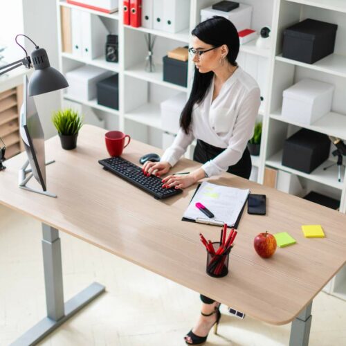 A woman stands at a desk working on a computer. The desk has office supplies, a red cup, an apple, and documents. Shelves with books, files, and boxes are in the background.
