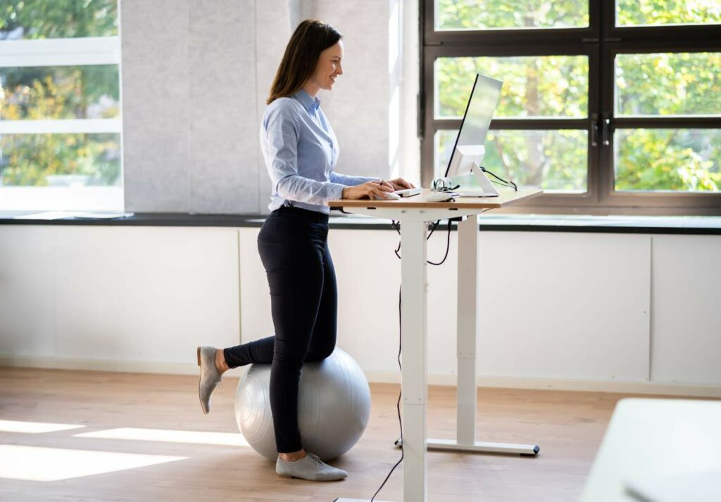 A woman uses a standing desk while sitting on an exercise ball in a bright, modern office space. She is looking at a computer screen and typing on the keyboard.