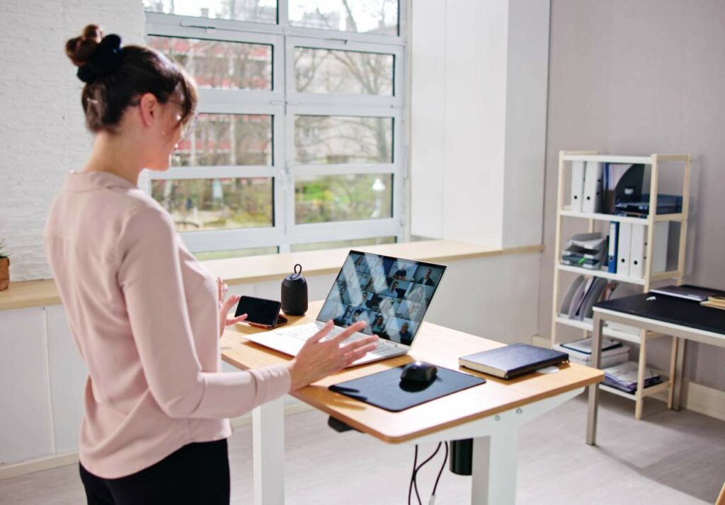 A person stands at a desk with a laptop, participating in a video conference. The desk also holds a phone, notebook, and other office supplies. Shelves and a large window are visible in the background.