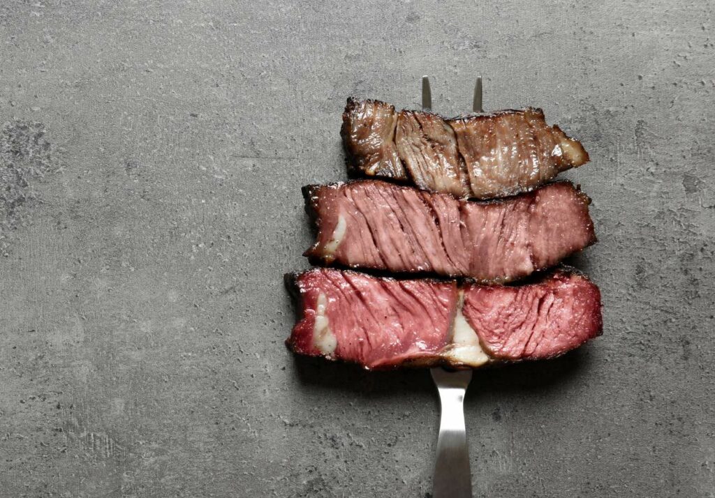 Slices of medium rare grilled steak on a fork, placed on a gray textured surface.