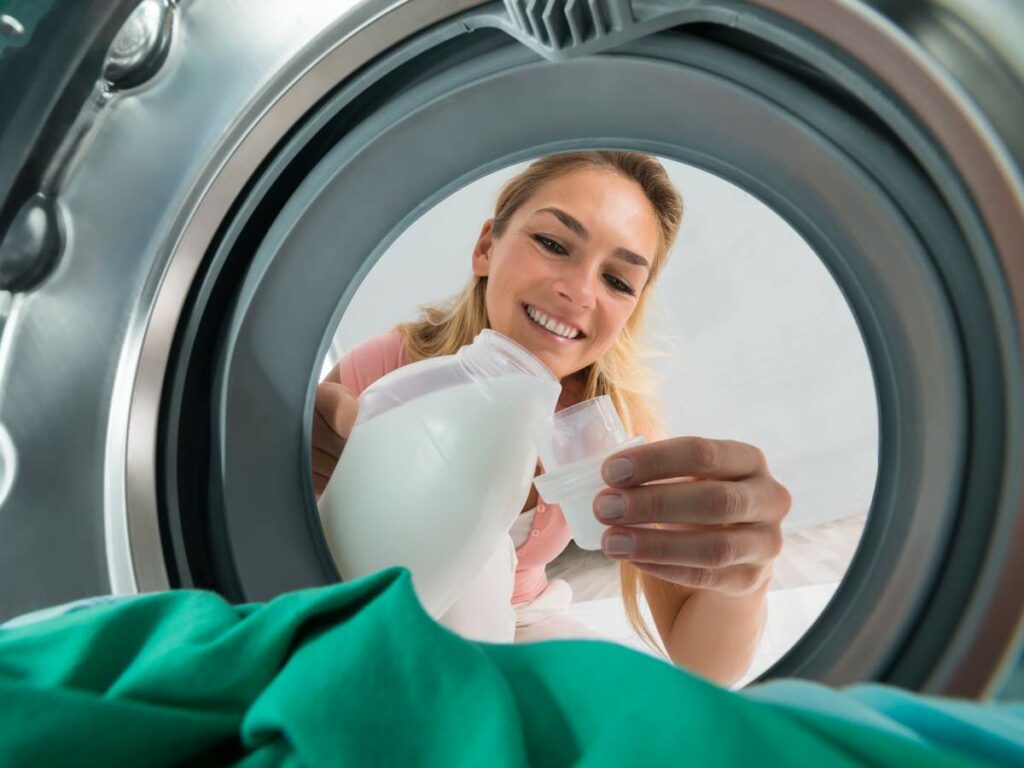 A woman smiles as she pours laundry detergent into a measuring cap, viewed from inside a washing machine.