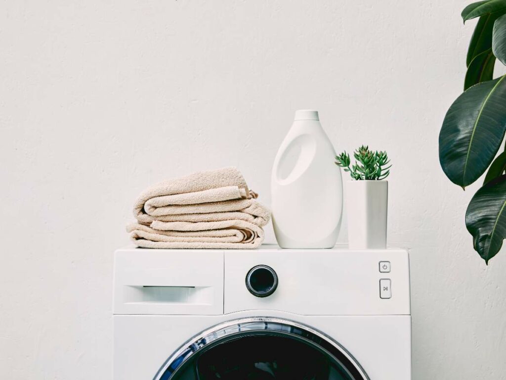 A washing machine with a folded stack of beige towels, a white detergent bottle, and a potted succulent on top. A green leafy plant is partially visible on the right side.