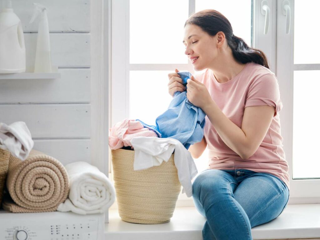 A woman in a pink shirt is sitting by a window holding a piece of laundry from a basket filled with clothes. Rolled towels are visible on a nearby shelf.