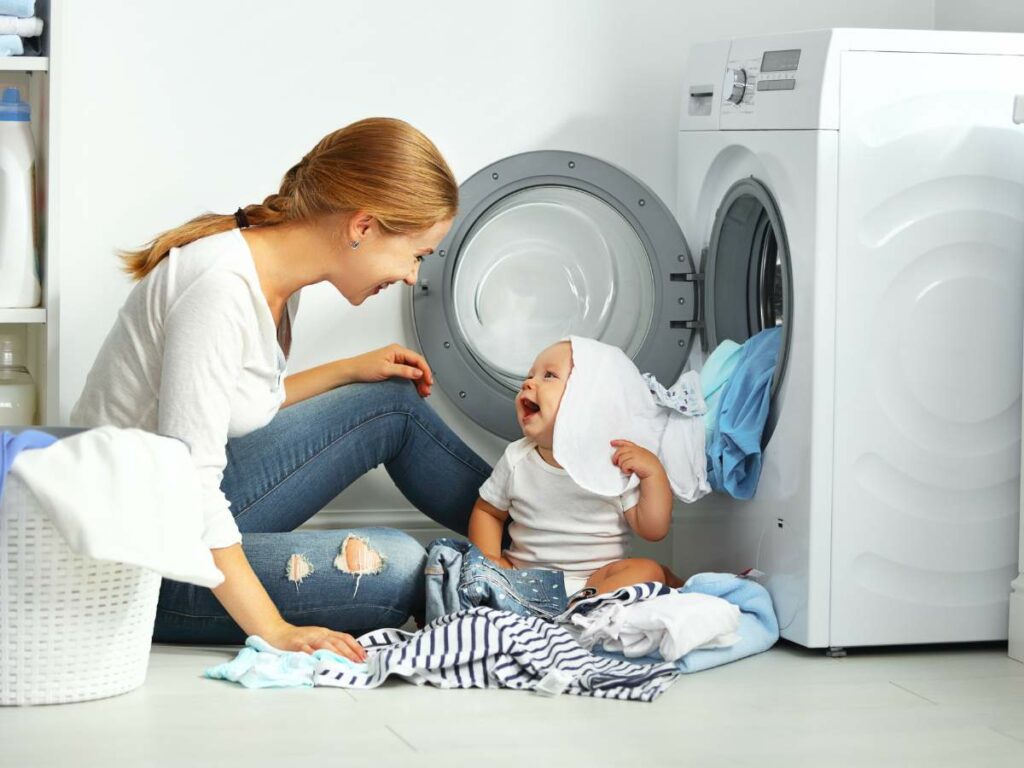 Woman and baby playing with laundry near a front-loading washing machine. The baby has a piece of cloth on their head, while the woman smiles at them. A laundry basket is in the foreground.