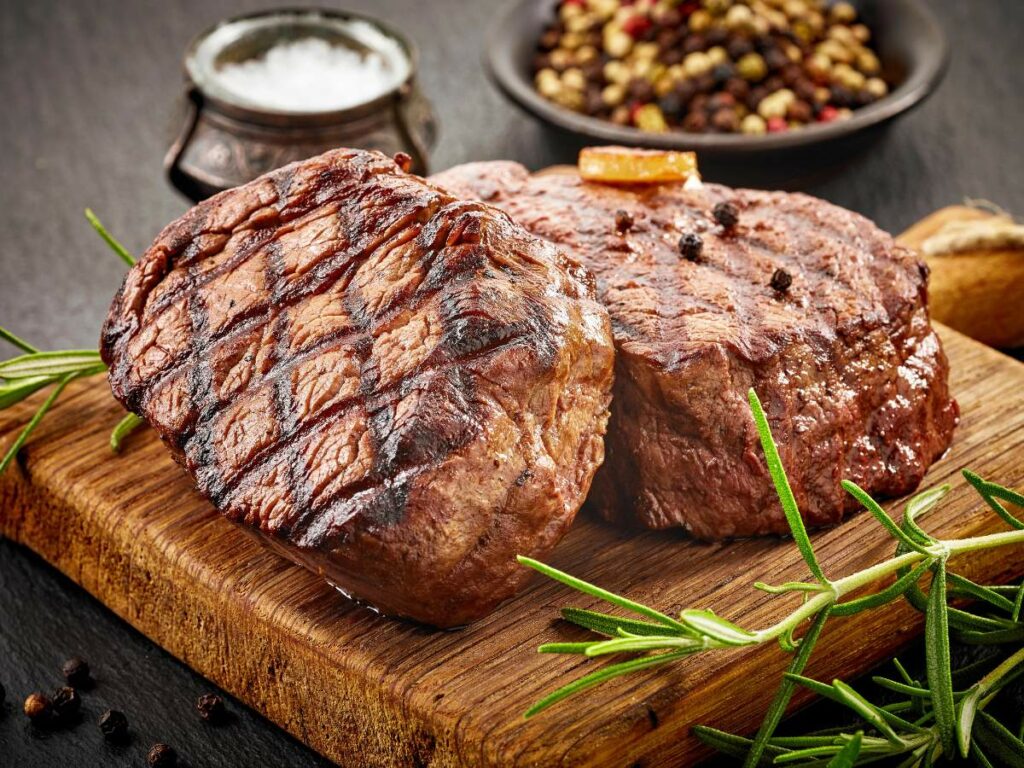Two grilled steaks on a wooden board, garnished with sprigs of rosemary, with a bowl of mixed spices and a container of salt in the background.