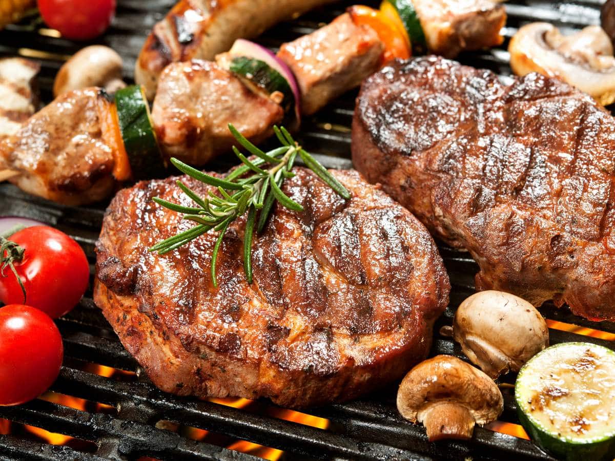 Close-up of various grilled meats, vegetables, and mushrooms on a barbecue grill, with tomatoes, zucchini, and rosemary on top.