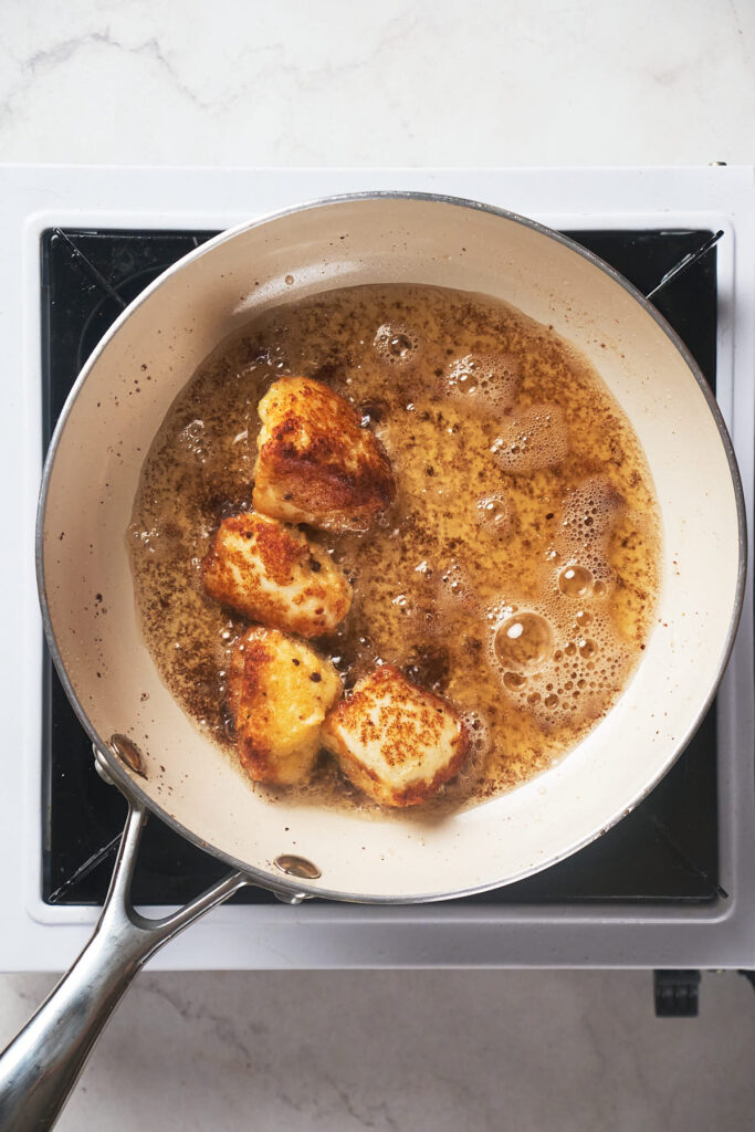 Four pieces of cheese curds are being pan-fried in lard in a white skillet on a stovetop.
