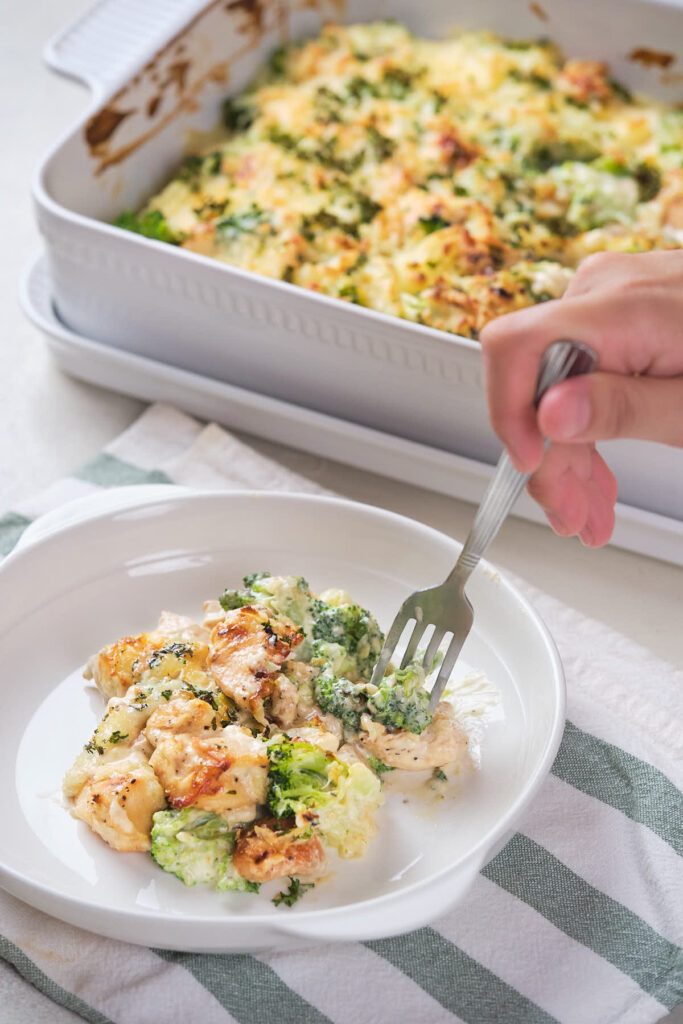 A hand holding a fork is about to eat a portion of chicken and broccoli casserole served on a small white plate.